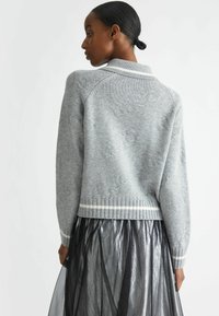 Maglione grigio a maglia con orlo a coste, strisce bianche e design del logo in rilievo. Indossato sopra una gonna in tulle nera trasparente, visto da dietro.