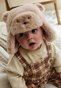 Purvinė šviesiai ruda meškutės kepurė su ausimis, apvaliu veidu ir minkšta tekstūra. Kūdikis dėvi ilgomis rankovėmis kremiškos spalvos marškinėlį ir rudą languotą kombinezoną.