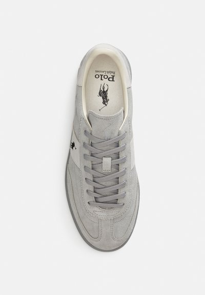 Grå mockasko med rundad tå, platta snören och en minimalistisk design. Har en krämfärgad rand och logotyp på sidan.