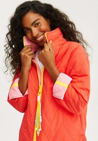 Poguněná bunda v jasném korálovém odstínu s růžovými manžetami a žlutým zipem. Obsahuje šňůrku na stažení s barevnými třásněmi pro úpravu.