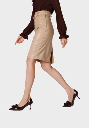 Donna che cammina indossando una gonna in pelle beige al ginocchio, una camicetta marrone a maniche lunghe e scarpe marrone scuro con tacco basso e fiocchi.