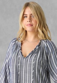 Blusa grigia a strisce con scollatura a V, spalle arricciate e maniche fluide. Il tessuto sembra leggero con una texture liscia e strisce bianche.