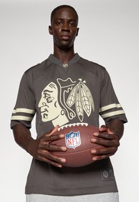 Jerseul sportiv de culoare gri închis, cu un grafic mare de nuanță bej reprezentând un șef american nativ și două dungi orizontale de culoare crem pe mâneci, ținând în mână un fotbal.