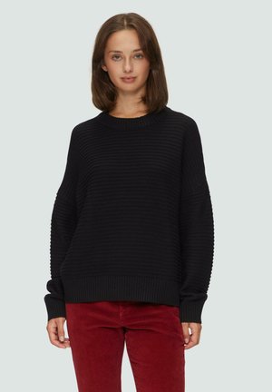 Schwarzer, gerippter Pullover mit Rundhalsausschnitt und weit geschnittenen Ärmeln; kombiniert mit roten Cordhosen. Die strukturierte Stoffe verleihen dem Design Tiefe.