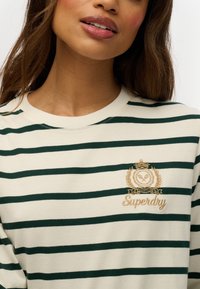 Crème-kleurige sweatshirt met groene horizontale strepen. Heeft een gouden geborduurd logo en kroontje op de borst. Zachte stoftextuur.