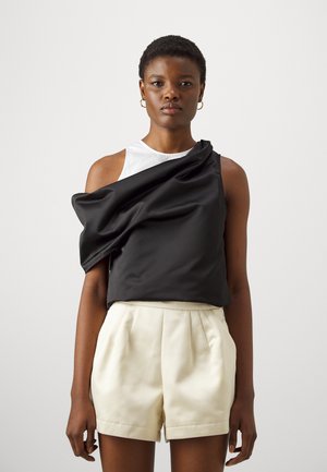Top senza maniche bianco e nero con un drappo di tessuto nero su una spalla, abbinato a pantaloni corti crema a vita alta. Texture liscia, design moderno.
