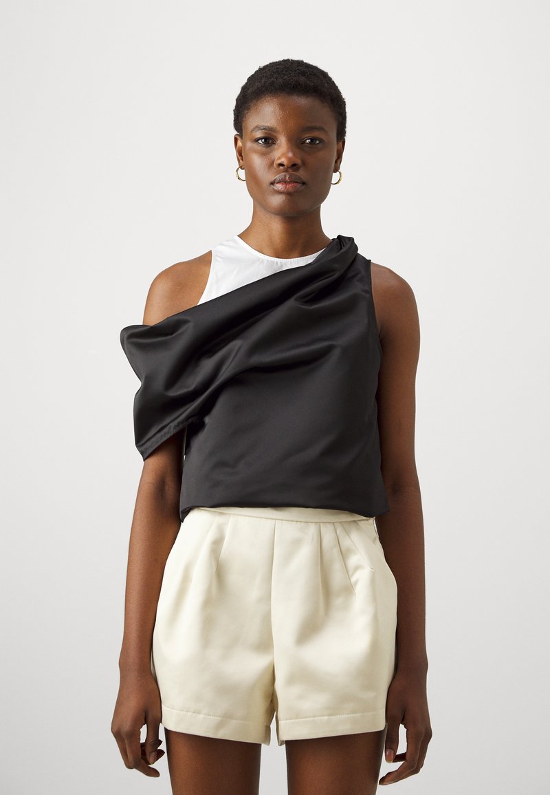 Top senza maniche bianco e nero con un drappo di tessuto nero su una spalla, abbinato a pantaloni corti crema a vita alta. Texture liscia, design moderno.