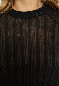 Melns adīts džemperis ar teksturētu, caurspīdīgu izskatu. Tajā ir vertikālas svītras un ribota, apaļa apkakle. Izceļas ar smalku mirdzumu.