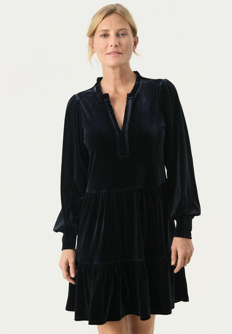 Rochie din catifea neagră cu un guler semi-deschis, mâneci lungi și puffate, și un tiv cu volane în straturi. Textură moale și strălucire subtilă.