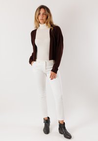 Cardigan marrone sopra un dolcevita bianco, abbinato a pantaloni bianchi. Stivaletti neri alla caviglia. Tessuto morbido, design aderente, silhouette lineare.