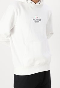 Bílá mikina s kapucí a přední kapsou, nastavitelná šňůrka na kapuci a červený a modrý text s logem "HILFIGER NEW YORK." Měkká tkanina.