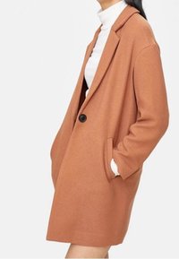 Frau trägt einen camel-farbenen Ein-Knopf-Mantel mit Seiten Taschen über einem weißen Rollkragenpullover, steht mit einer Hand in der Tasche.