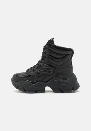 Schwarzer Knöchelstiefel mit glänzendem Synthetik-Obermaterial, gepolstertem Kragen, schnürbarem Vorderteil und einer robusten Gummisohle mit strukturierten Akzenten.