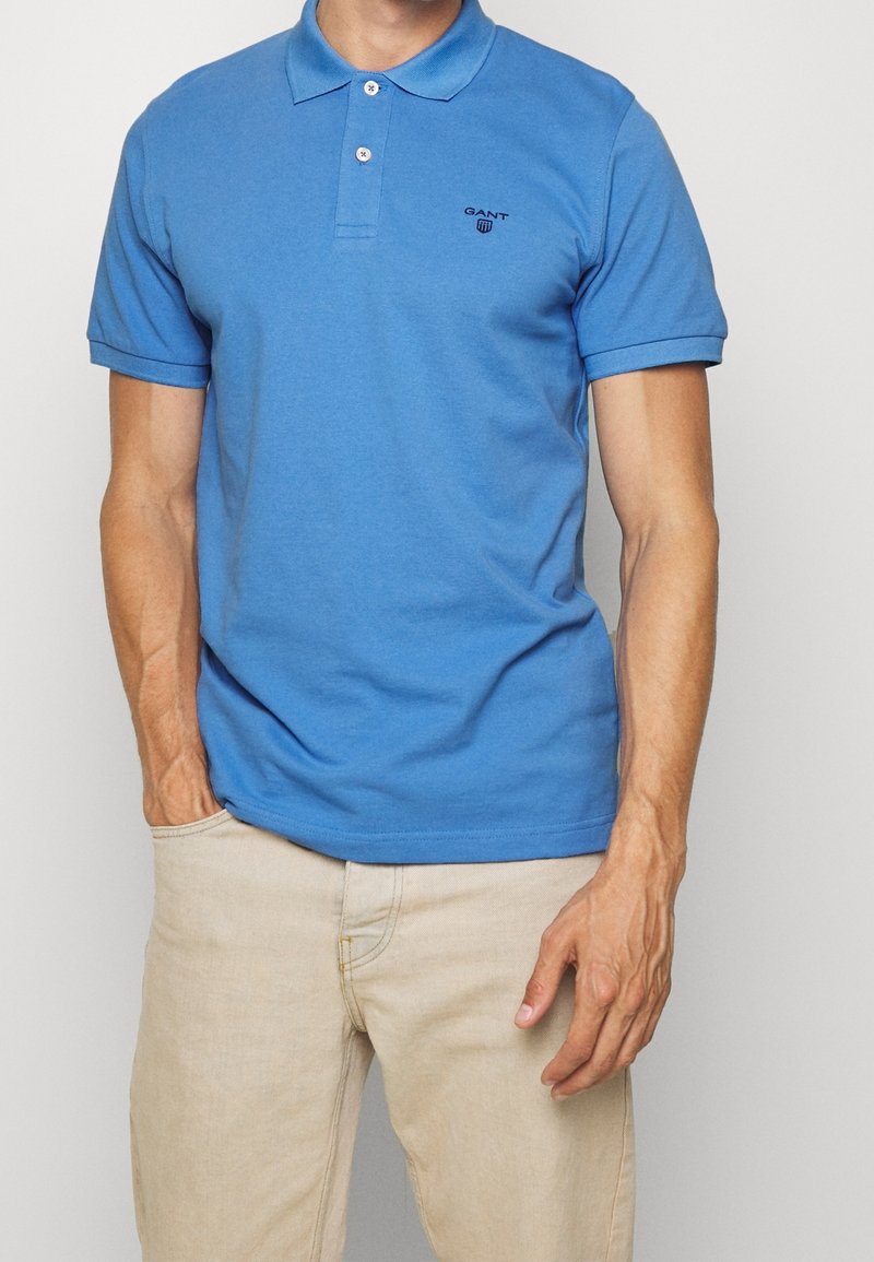 Světle modrá polo košile vyrobená z bavlny, se klasikým límečkem, krátkými rukávy, dvěma knoflíky a malým černým logem na levé hrudi.