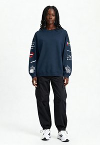 Donna con capelli intrecciati che indossa una felpa blu navy con stampa colorata sulle maniche, pantaloni cargo neri e scarpe da ginnastica bianche, in piedi su uno sfondo bianco.