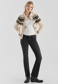 Strickjacke in Creme mit schwarzen geometrischen Mustern, kombiniert mit dunklen Jeans und schwarzen Stiefeletten mit silbernen Akzenten.