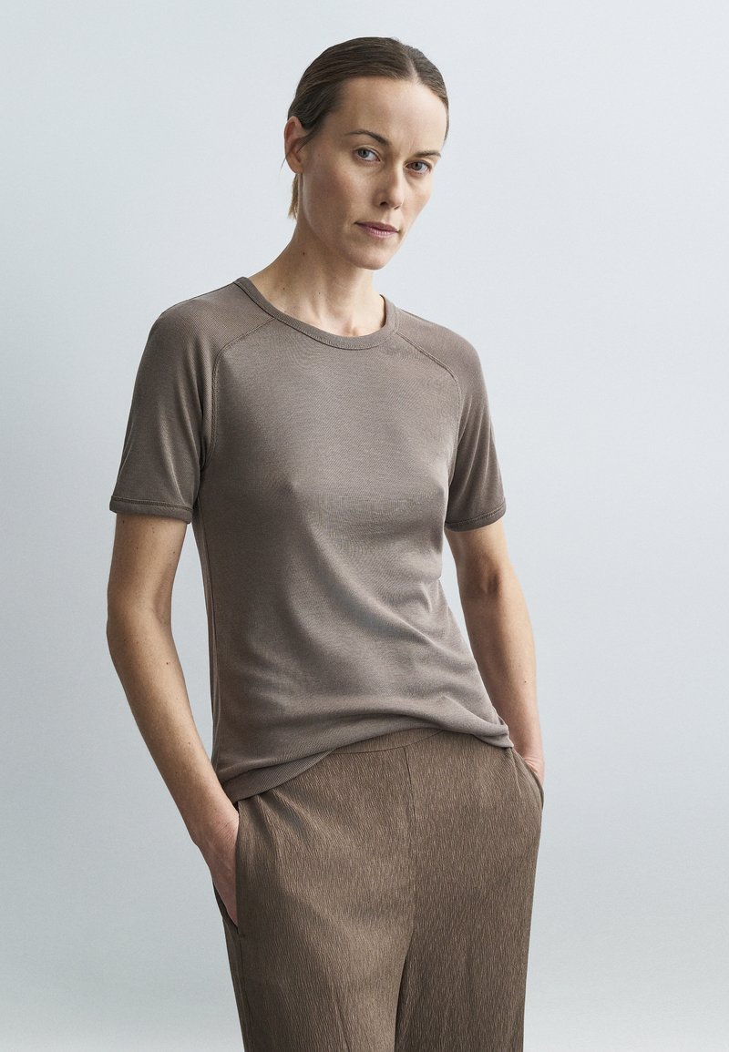 Sieviete ar gaišu ādu un atpakaļ savilktiem matiem, ģērbta īsroku brūnā topā un atbilstošās struktūrētās biksēs, rokas kabatās, neitrāls fons.