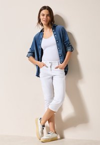 Chemise en denim, débardeur blanc, pantalons blancs raccourcis et baskets légères. La chemise a des manches retroussées et les pantalons présentent des coutures discrètes.