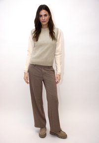 Pull beige clair en tricot porté sur un top crème à manches longues, associé à un pantalon large marron à motifs et des chaussures beige clair.