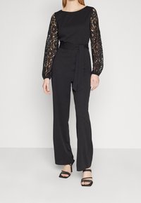 Svart jumpsuit med långa spetsärmar, figursydd kropp, vida byxor och matchande bälte. Slät tygtextur med intrikata spetsdetaljer.