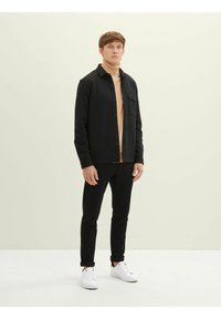 Chemise noire à boutons sur un col roulé beige, associée à un pantalon noir et des baskets blanches. Tissu lisse, coupe ajustée et longueur moyenne.