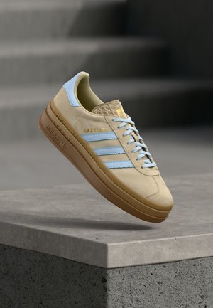 Bēša zamšādas sporta apavs ar gaiši ziliem akcentiem un trim svītrām. Tās ir ar fakturētu papēdi, paceltu zolī un uz sāniem iespiestu logotipu.