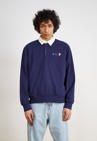 Mörkblå sweatshirt med vit krage och frambroderad logotyp i flera färger. Lätt passform och mjuk textur, kombinerad med ljusa denimjeans.