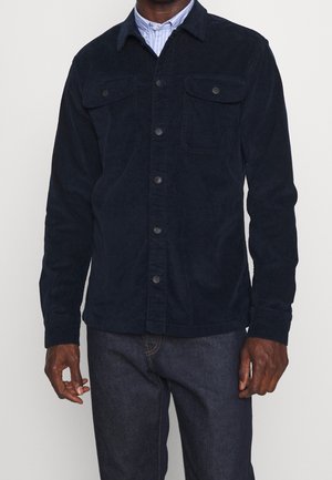 Homme portant une veste boutonnée en velours côtelé bleu marine foncé sur une chemise à col rayée bleu clair et un jean bleu foncé.
