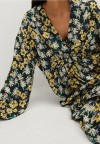 Kukkamekko mustasta kankaasta, jossa on keltaisia ja valkoisia kukkia, pitkät hihat, V-pääntie, kootut vyötärö ja virtaava hame.