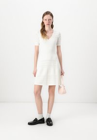 White dress with textured pattern, short sleeves, and a V-neck. Paired with black shoes, white socks, and holding a small pink handbag.