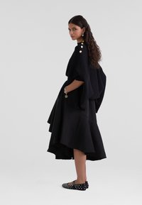 Čierny oversized pletený top s rebrovaním a zlatými gombíkovými doplnkami, skombinovaný s vrstvenou čiernou sukňou a čiernymi balerínami s bielymi bodkami.