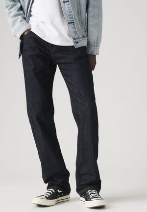 Personne portant un jean droit foncé, des baskets noires et blanches, un t-shirt blanc et une veste en denim bleu clair avec une main dans la poche.
