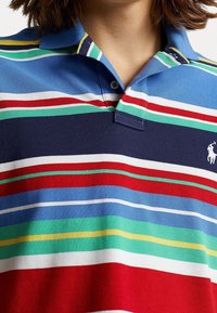 Pikétröja med horisontella ränder i blått, marinblått, rött, grönt och vitt. Mjuk bomullstyg med krage och knäppning med två knappar.