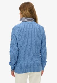 Jasnoniebieski dziany sweter z wzorem warkocza, ze ściągaczami na mankietach i u dołu, noszony na koszuli z paskowanym kołnierzykiem, prezentujący teksturowany design.