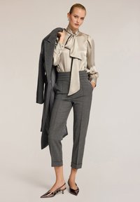 Pantaloni grigi a quadri con vita alta, abbinati a una leggera blusa di seta con un fiocco. Un cappotto grigio scuro appoggiato sulla spalla e scarpe con tacco metalliche.