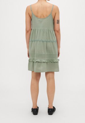 Femme portant une robe sans manches vert sauge avec des volants et des détails en dentelle, des chaussures noires et un tatouage géométrique sur son bras gauche supérieur, debout de dos.