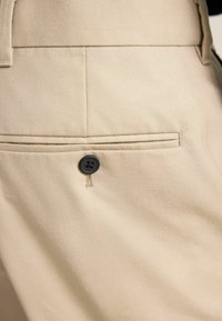 Beige Hose mit einer Gesäßtasche mit horizontalem Schlitz und einem schwarzen Knopfverschluss. Der Stoff hat eine glatte, strukturierte Oberfläche.