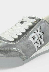 Baskets argentées avec lacets blancs et "DKNY" brodé en blanc et noir sur le côté, montrant des coutures détaillées et un matériau texturé.