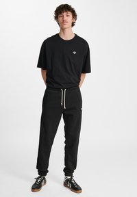 Schwarzes T-Shirt mit einem kleinen weißen Herzlogo auf der Brust, kombiniert mit schwarzen Sweatpants mit elastischem Bund und Kordelzug.