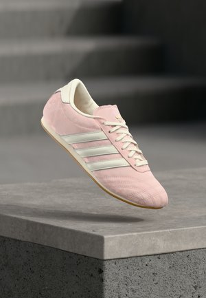 Ljusrosa Adidas-sneaker med vita ränder och snören, visas i luften ovanför en betongkant med suddiga trappsteg i bakgrunden.