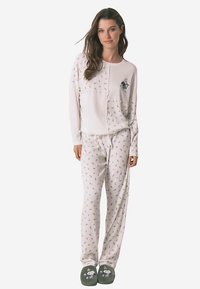 Pyjama set met lange mouwen in lichtcrème, bestaande uit een getextureerd shirt en een broek met patroon. Heeft een grafisch accent en zachte groene slippers.