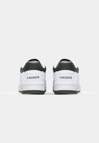 Adidași albi cu accente negre, cu un superior din piele netedă, guler căptușit și talpă din cauciuc. Logo-ul "LACOSTE" pe toc.