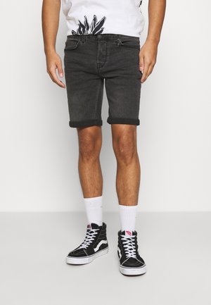 Hombre que lleva pantalones cortos de mezclilla oscuros enrollados, calcetines blancos a media pantorrilla, zapatillas negras de caña alta y una camisa blanca con un diseño floral negro.