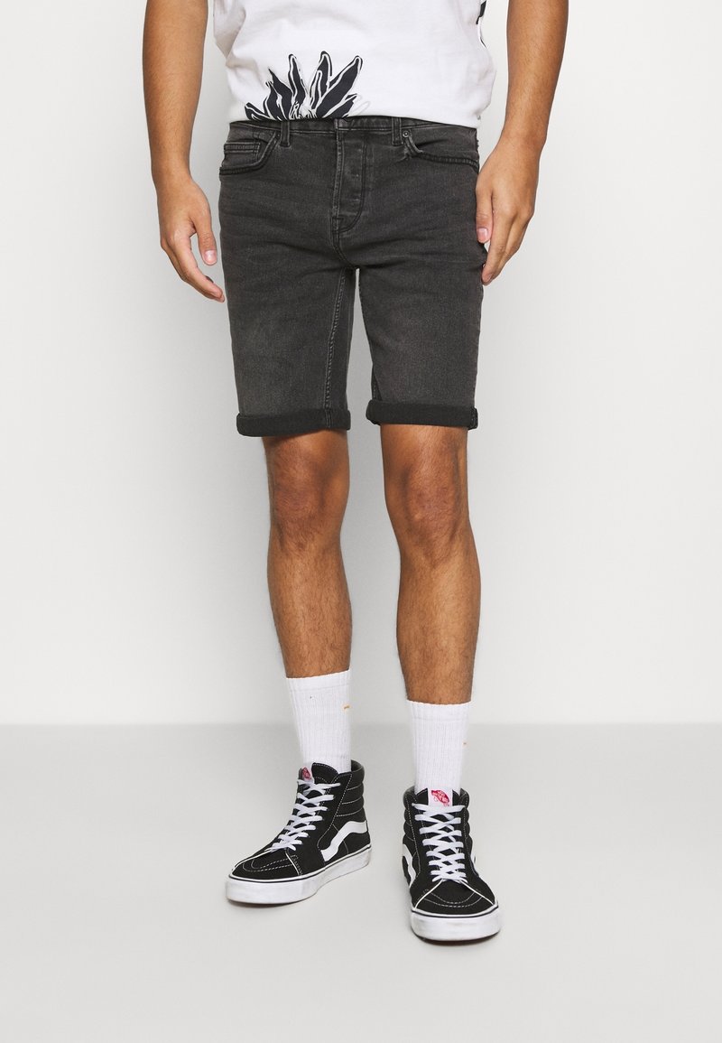 Hombre que lleva pantalones cortos de mezclilla oscuros enrollados, calcetines blancos a media pantorrilla, zapatillas negras de caña alta y una camisa blanca con un diseño floral negro.