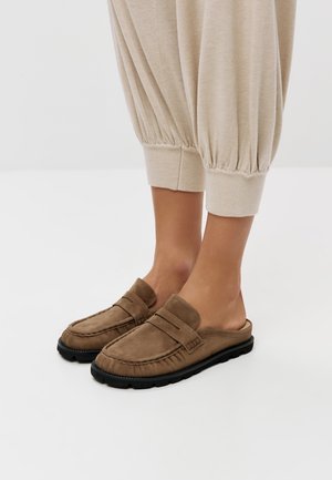 Piedi che indossano mocassini taupe in camoscio senza lacci con suole nere, abbinati a pantaloni beige risvoltati su sfondo bianco.