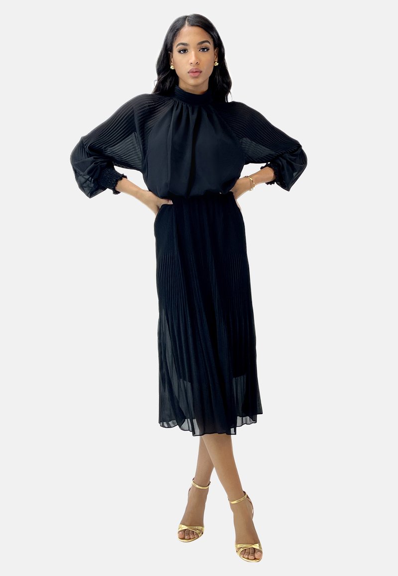 Vestido midi plisado negro con un cuello alto y mangas abullonadas. Presenta un tejido transparente, una cinturilla elástica y sandalias con detalles en dorado.