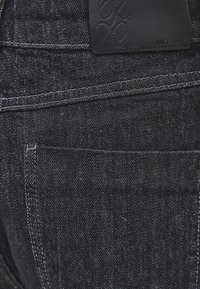 Nærbillede af baglommen på mørkeblå denimjeans med detaljerede syninger og en sort læbeetiket syet fast ved linningen.