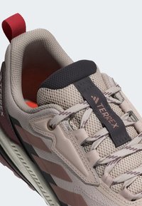 Chaussure de trail Terrex beige clair et rose avec tige en mesh, col rembourré, lacets à motifs et languette de tirage noire marquée.