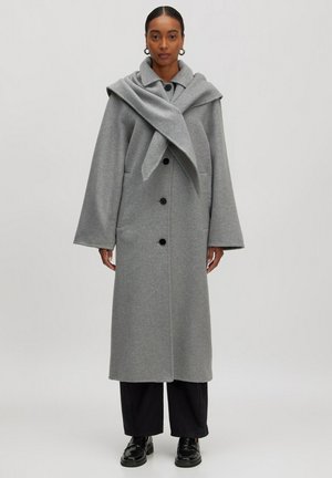 Lange grijze wollen blend jas met een sjawl-kraag, voorzien van een knoopsluiting en zijzakken, gecombineerd met zwarte wijde broek en schoenen.