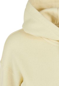 Sweat à capuche beige clair montrant la partie supérieure de l'épaule et une grande capuche repliée avec des détails de couture visibles.